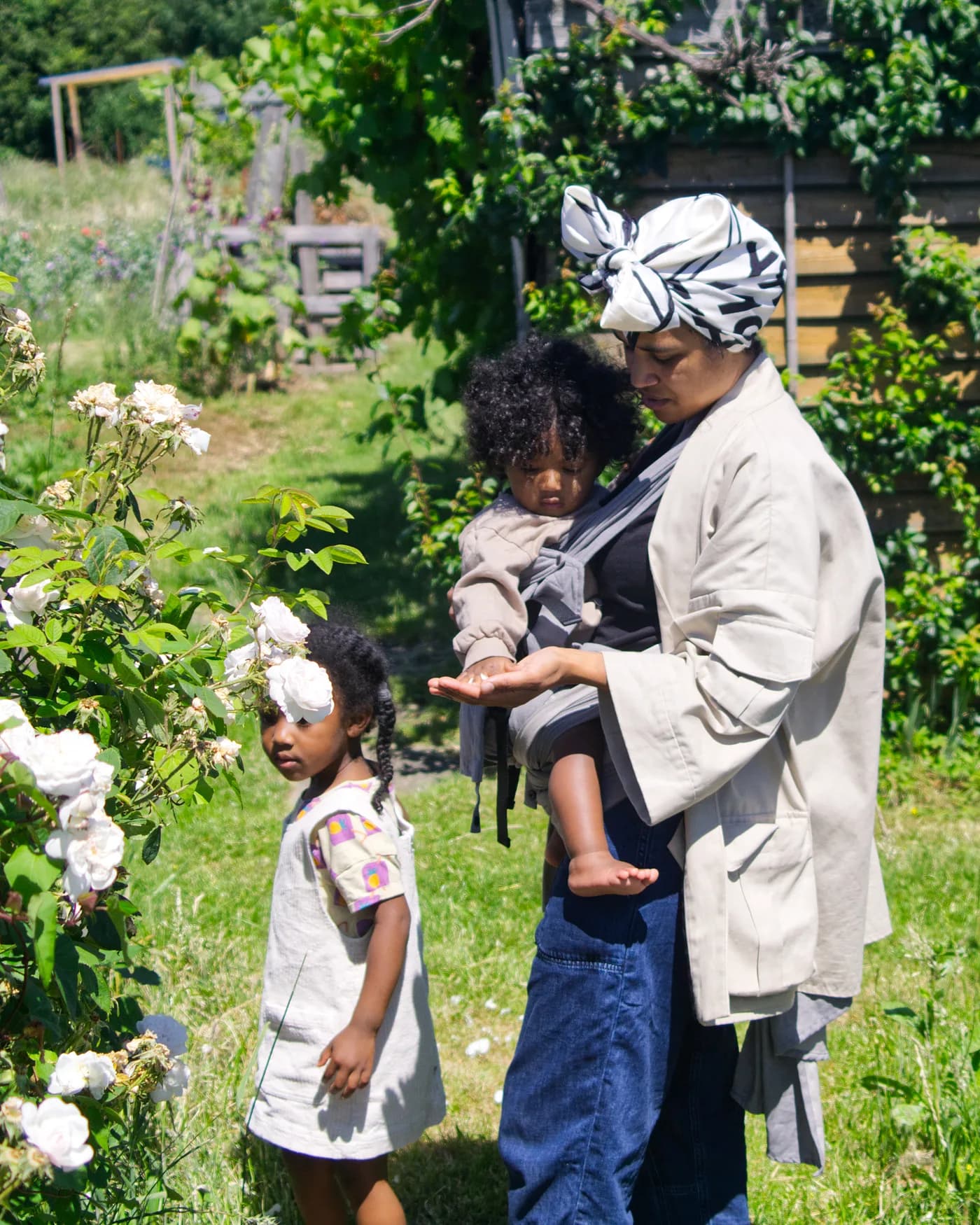 A parent in a garden carrying her toddler in the Mbereko, an older child beside her